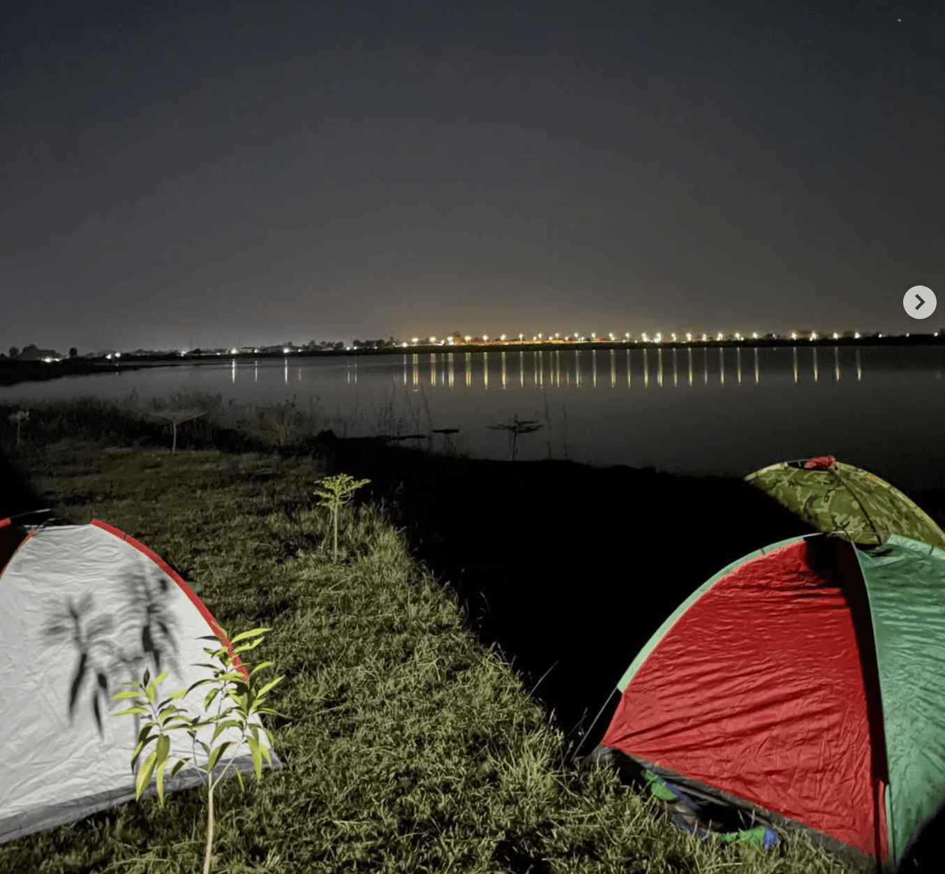 Tents pitched by a waterfront at night with city lights reflecting on the water