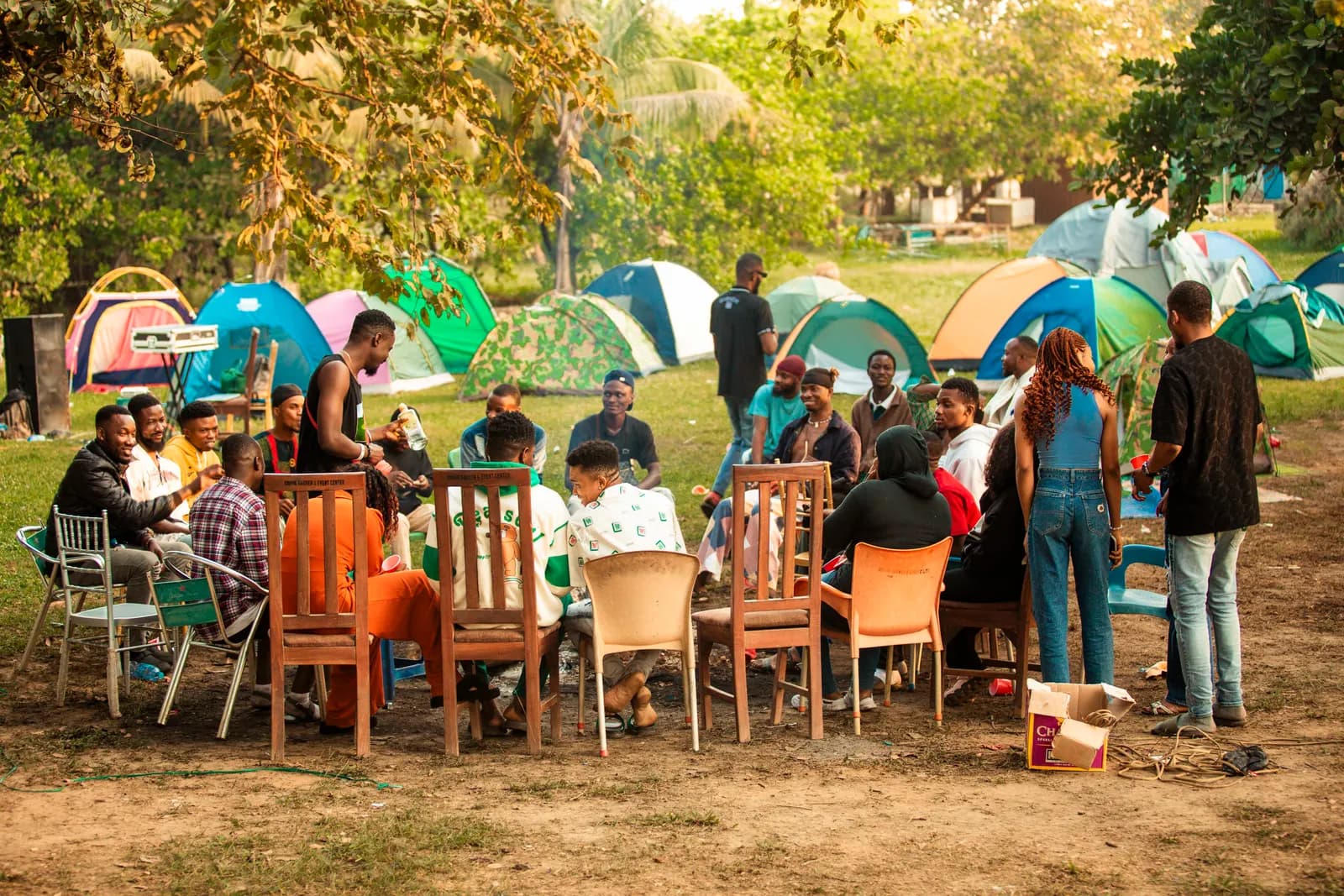 Community members sitting in a wide circle of chairs on the grass, with colourful tents pitched behind them