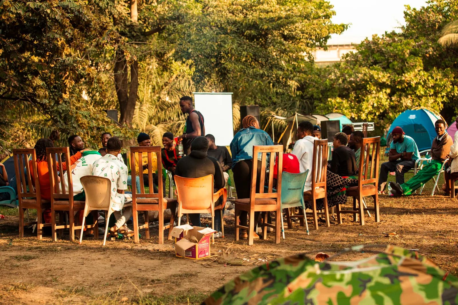 Members seated on wooden chairs in a circle under the trees