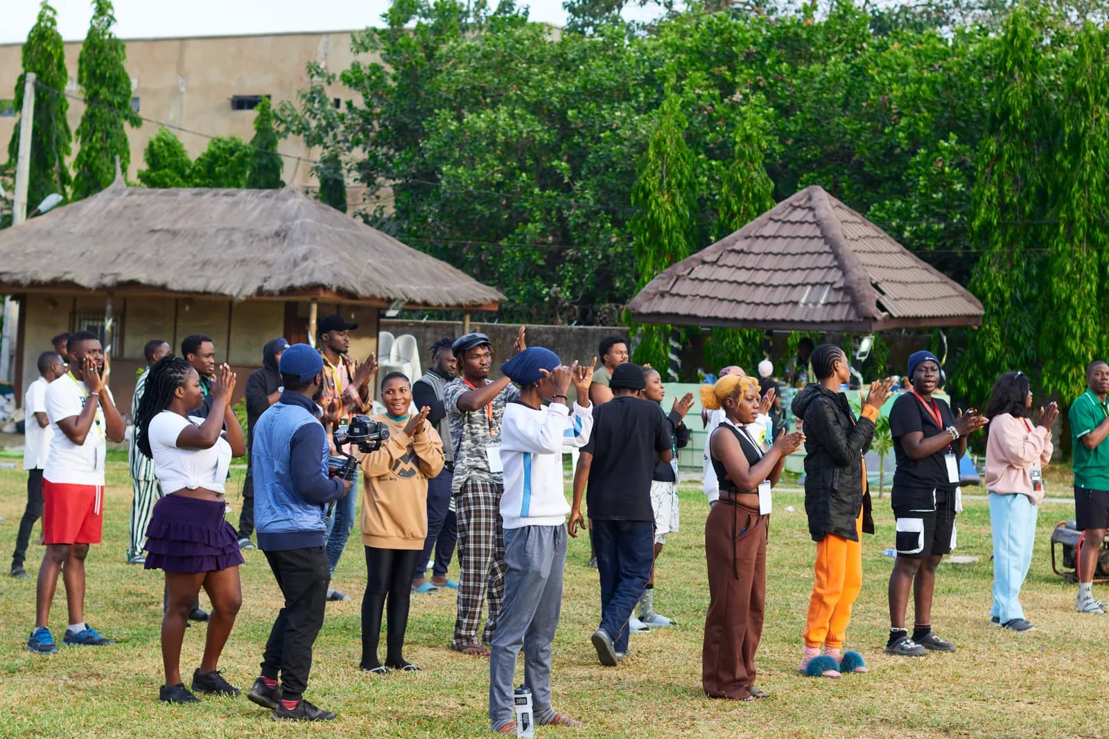 A large group of community members gathered on a lawn beside thatched huts, joining in a group activity