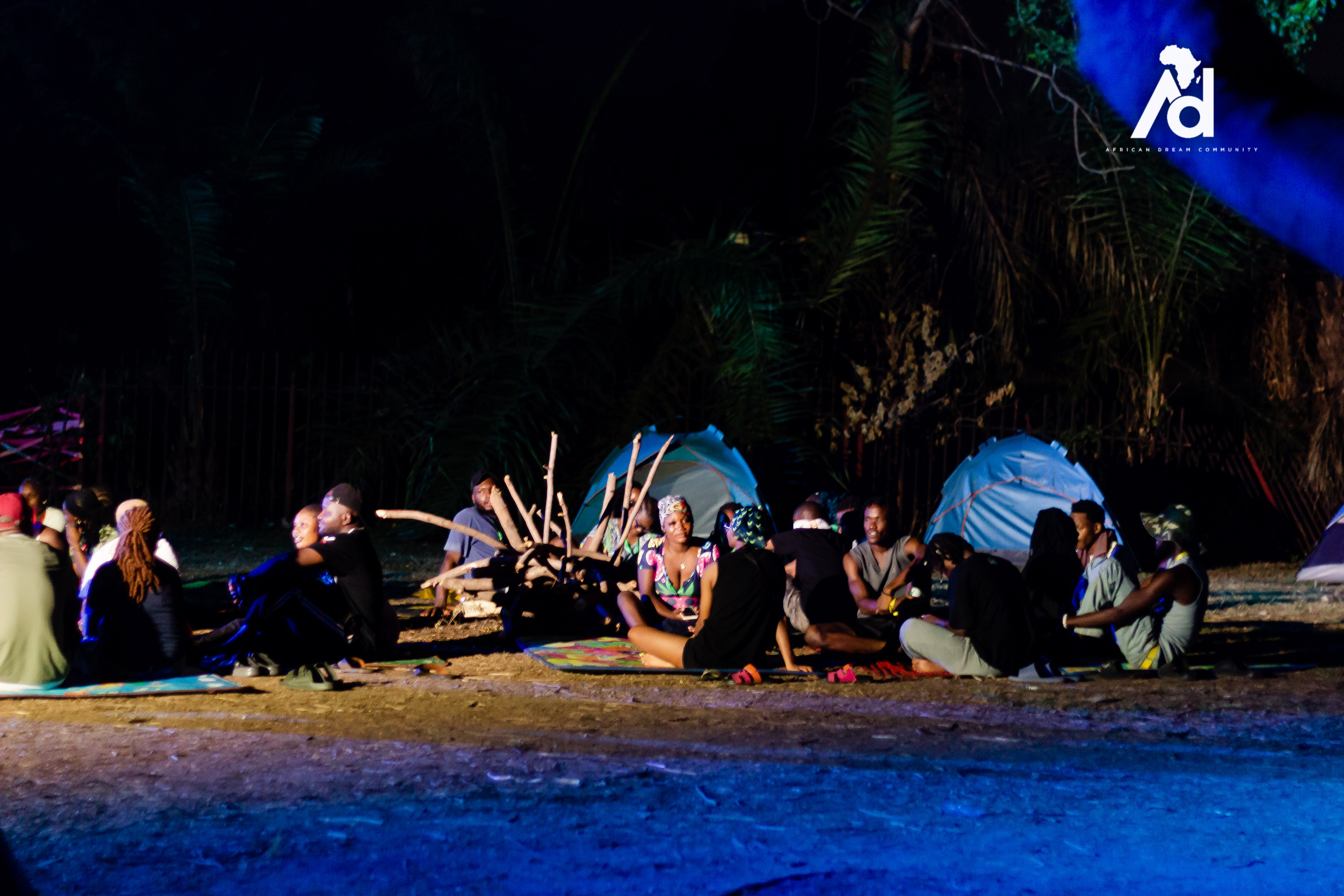 Group of people enjoying a nighttime camping experience around a campfire with blue tents in the background
